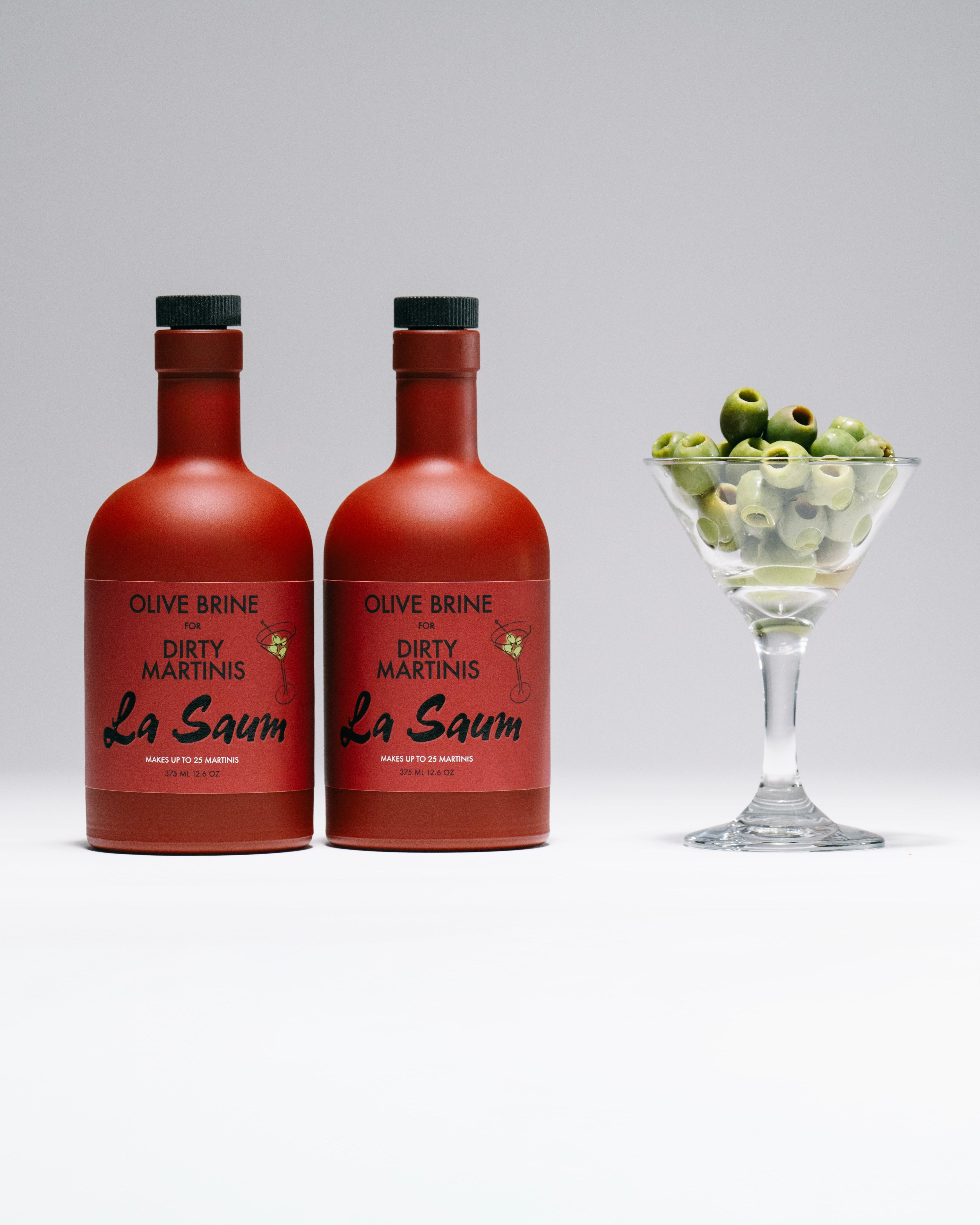 Two bottles of Olive Brine Dirty Martini La Saum with a glass of cocktail and olives on a white background
