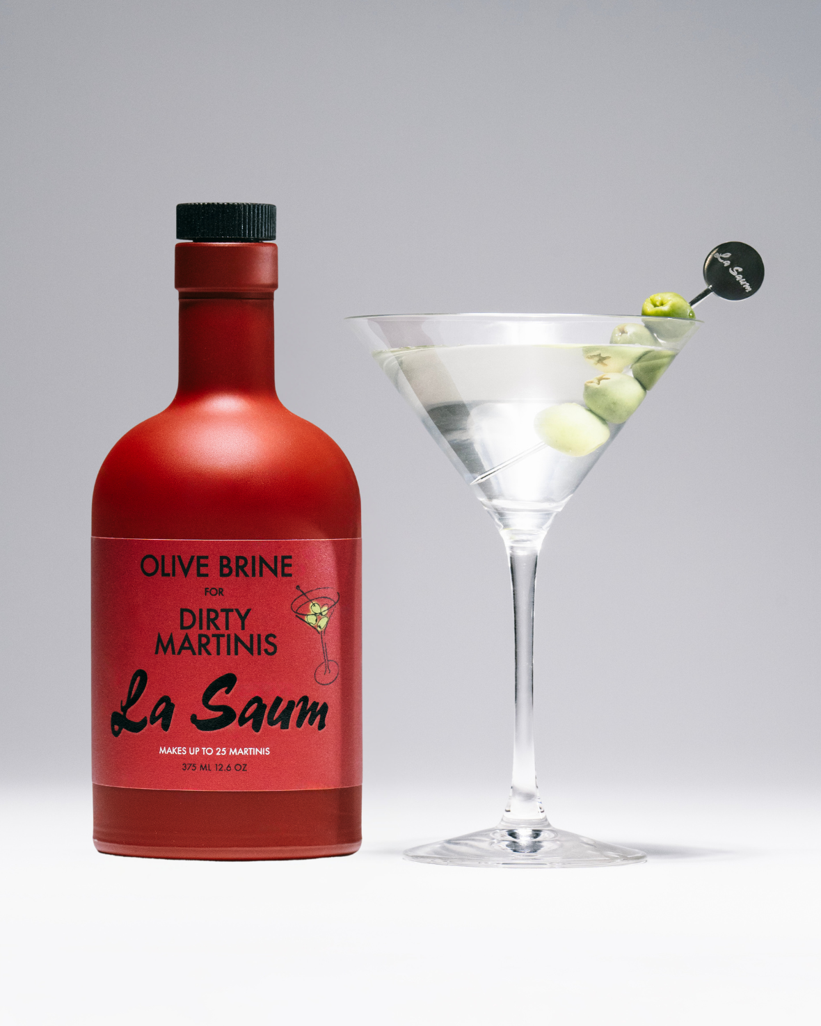 Red bottle labeled 'Olive Brine Dirty Martinis La Saum' next to a martini glass with a olive on a gray background