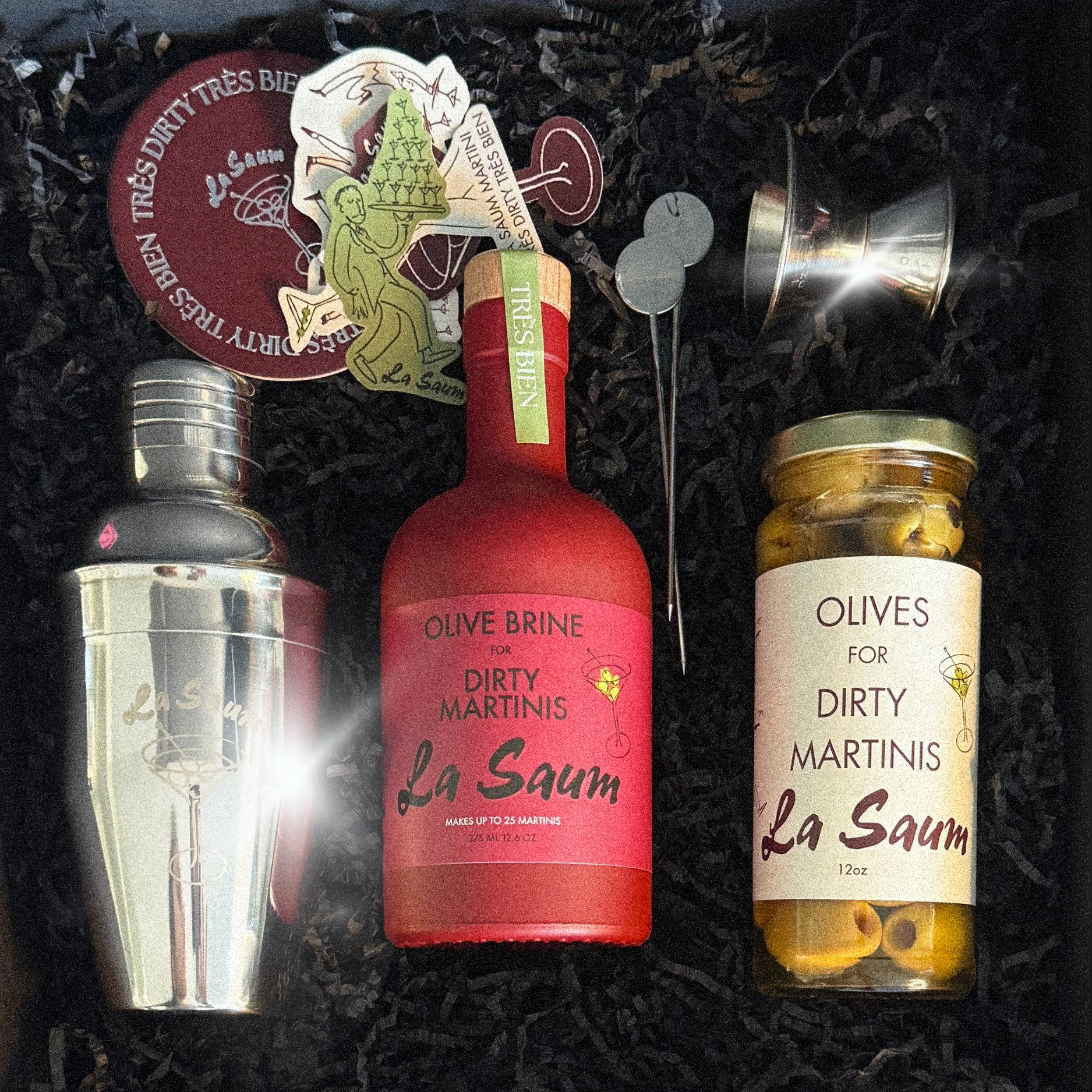 La Saum olive brine and olive jar with cocktail shaker and garnish on a dark background
