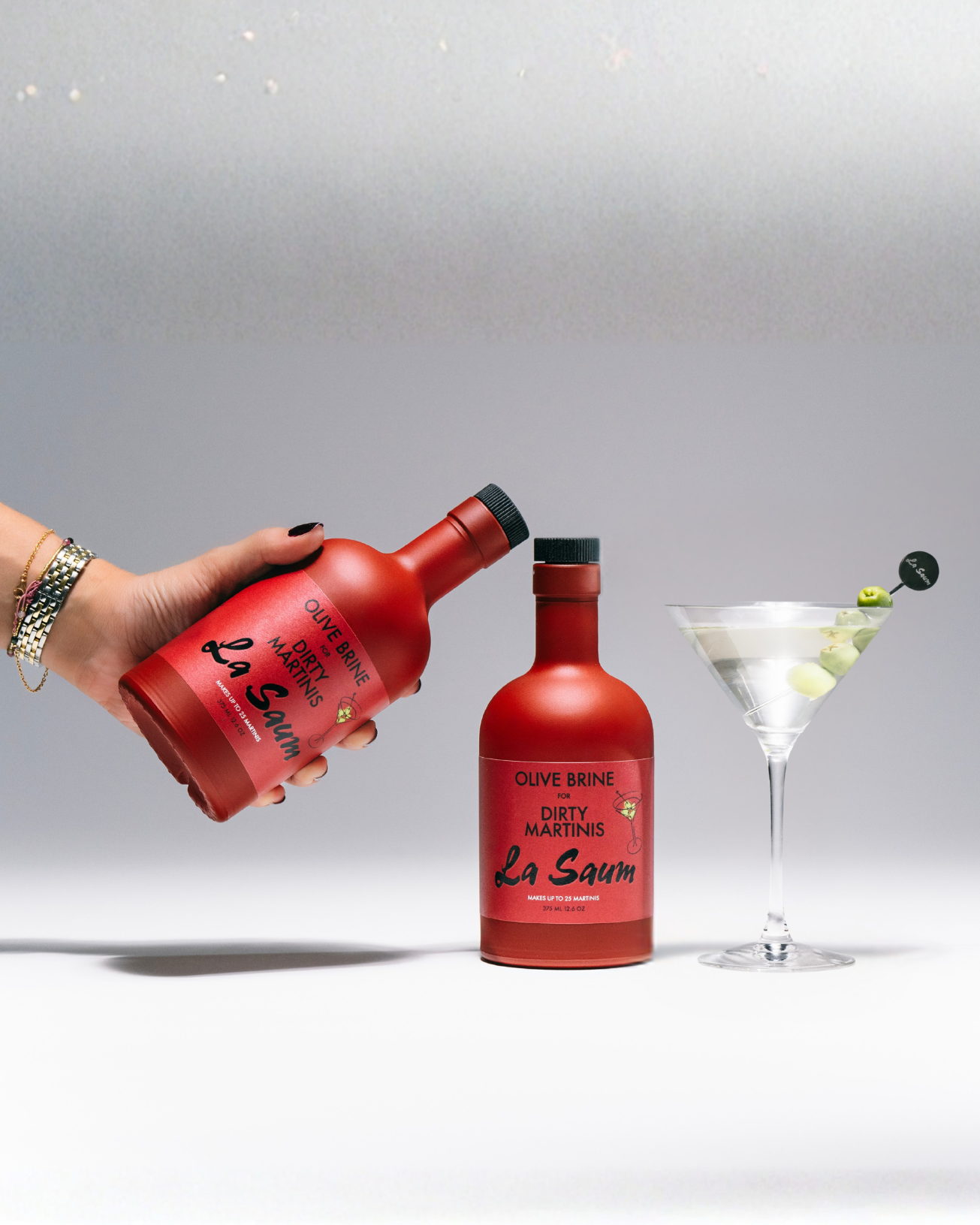 Red bottle labeled 'La Saum' held by a hand, with another bottle and a cocktail in the background.