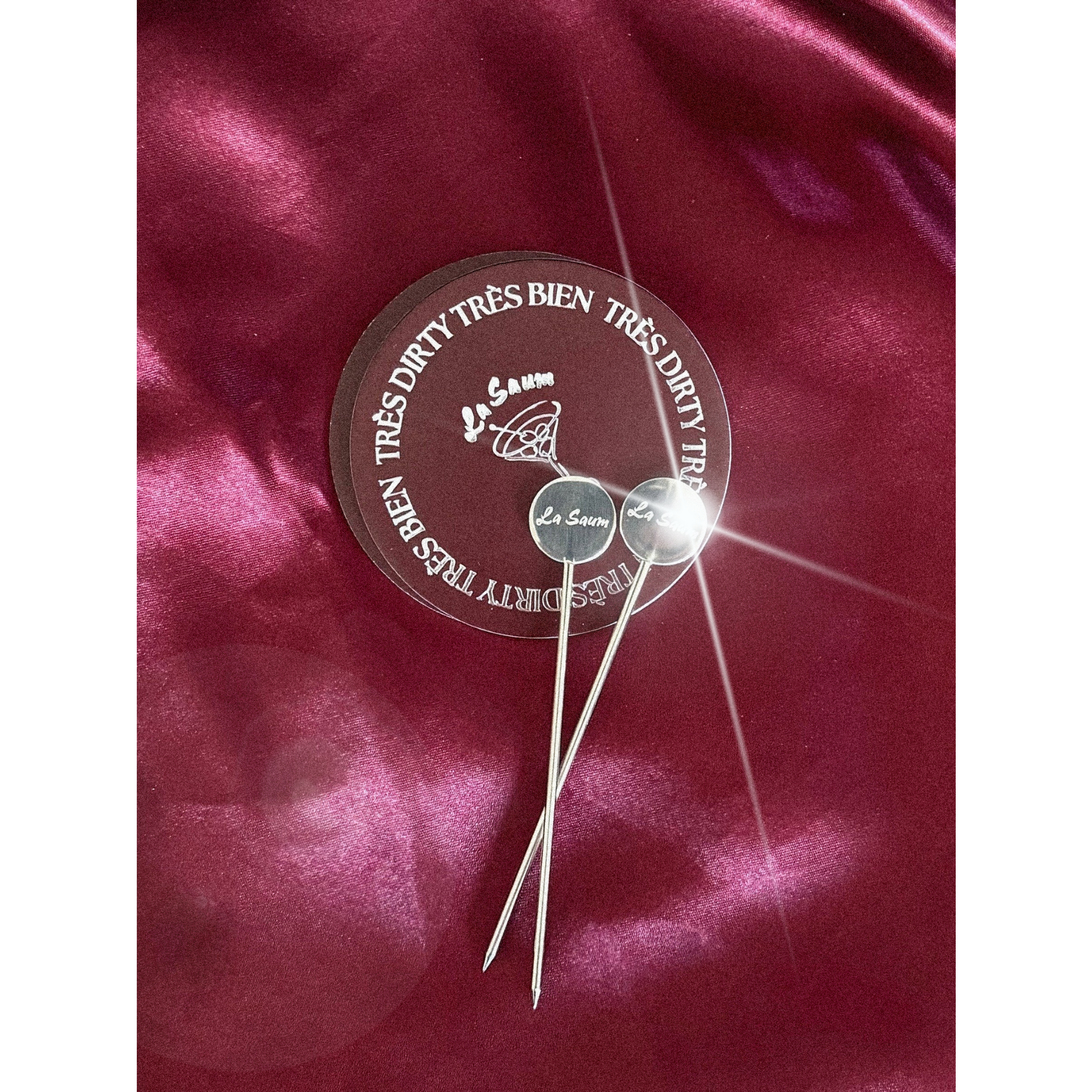Two silver cocktail picks with a circular la saum olive brine branded label on a red satin background