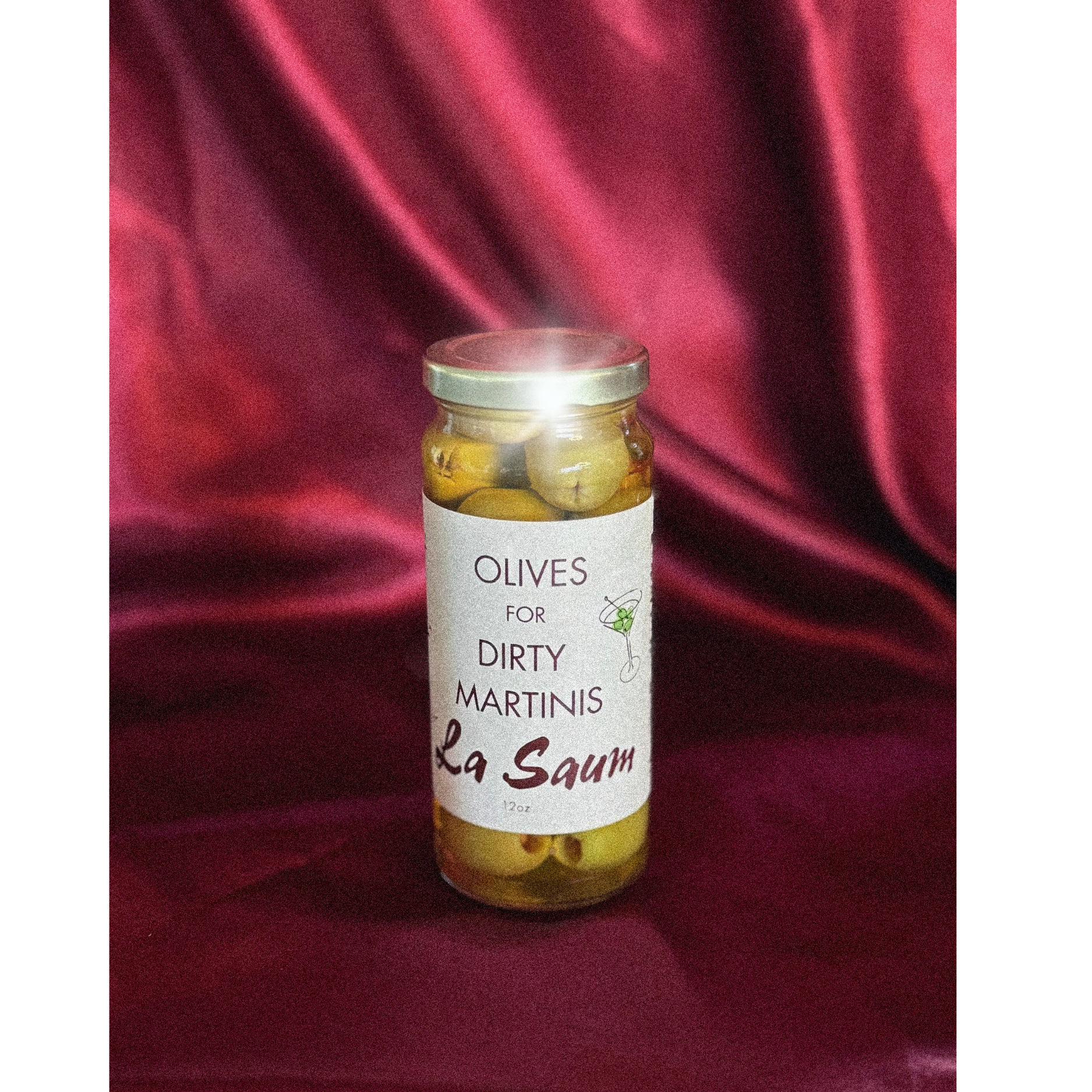 Jar of olives labeled 'Olives for Dirty Martinis La Saum' on a red fabric background